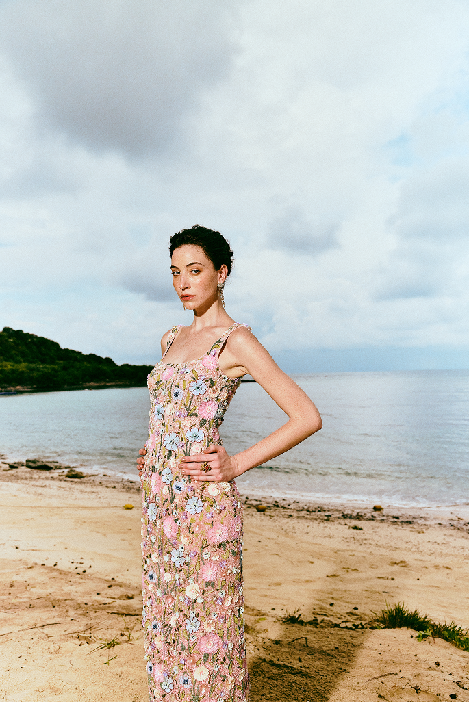 A woman stands on a sandy beach wearing a sleeveless, floor-length floral dress with pink, peach, and blue tones. The background shows the ocean, cloudy sky, and shoreline.