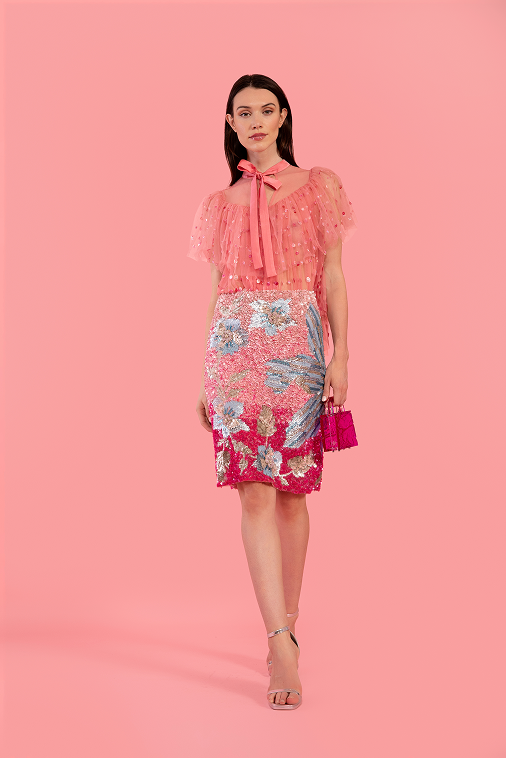 A model stands against a solid pink studio background wearing a sheer pink sequined top with a bow tie-neck and a pink floral lace midi skirt, holding a small pink handbag.