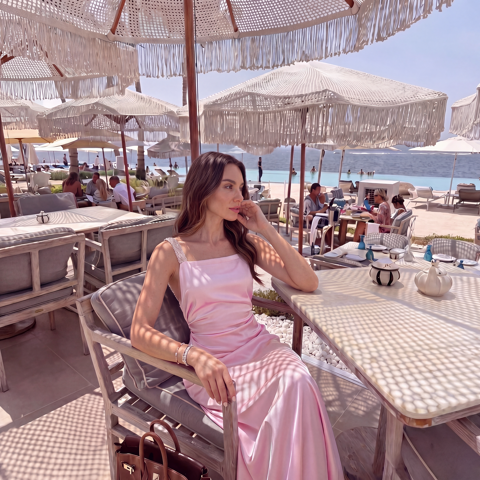 Woman in a pink dress sitting under a straw umbrella at a beachside cafe.