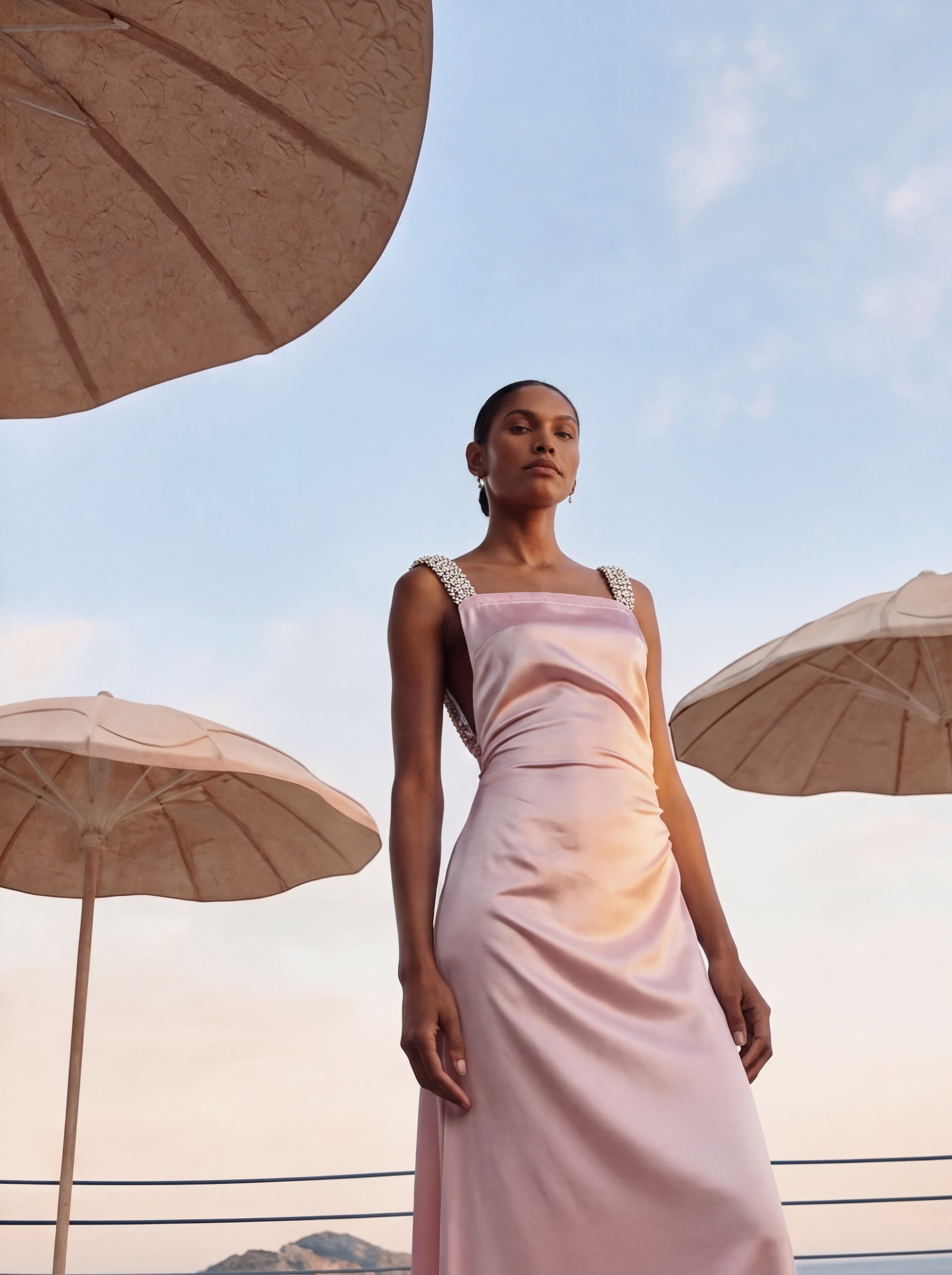 Woman in a pink dress standing under beige umbrellas with a scenic background
