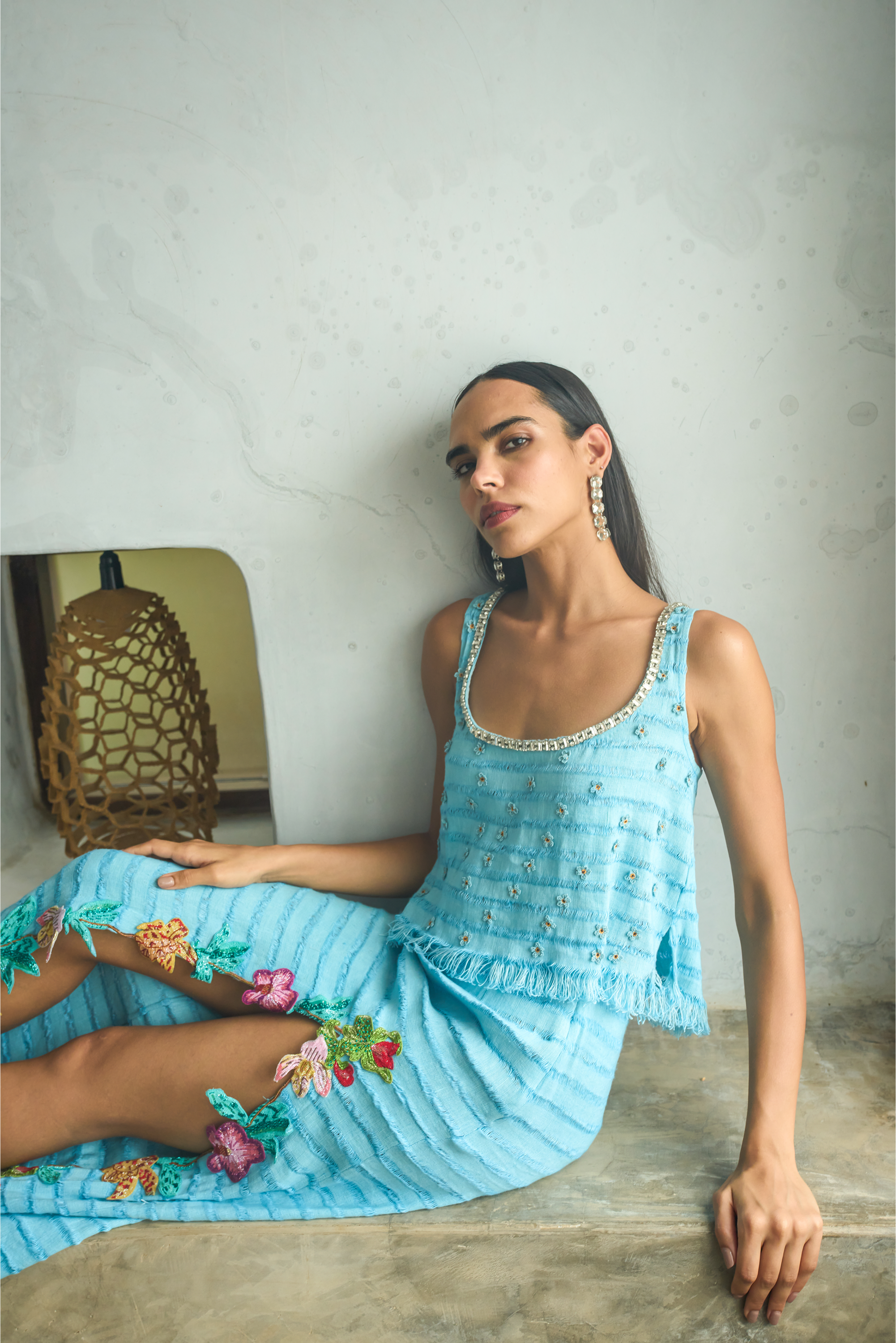Woman in a blue embroidered dress sitting on a wooden bench against a plain wall.