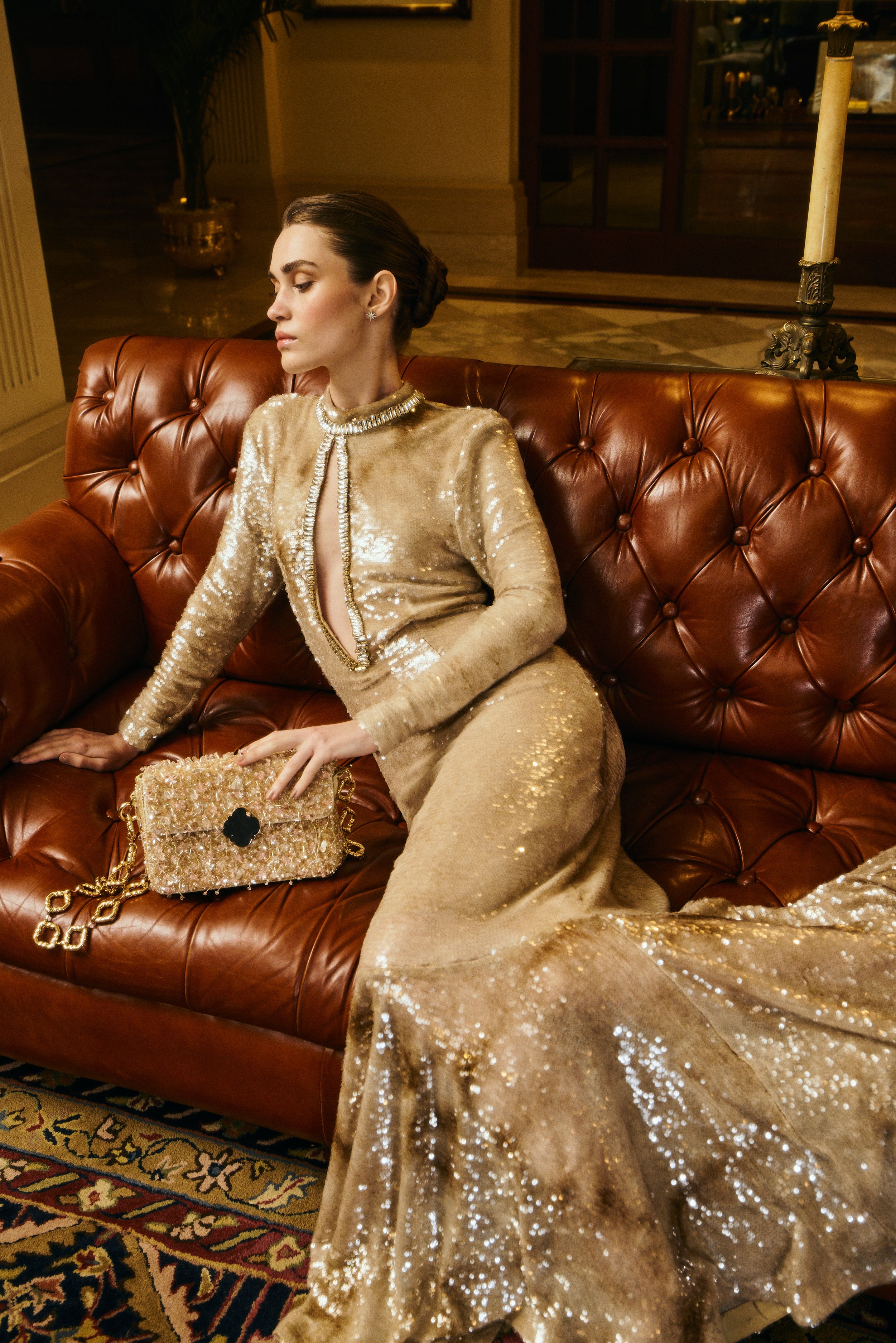 Woman in a gold sequin dress sitting on a brown leather couch in an elegant room.