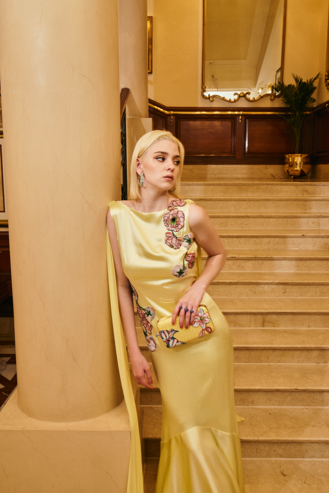 Woman in a yellow dress with floral patterns leaning against a column in an elegant interior setting.