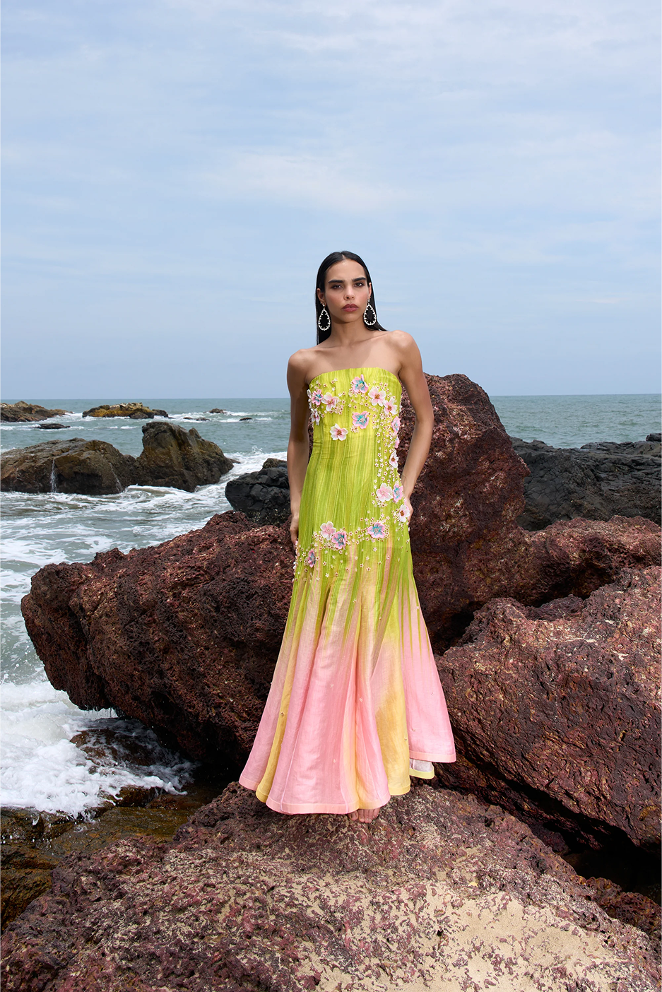 Woman in a strapless dress with floral patterns standing on rocks by the ocean.