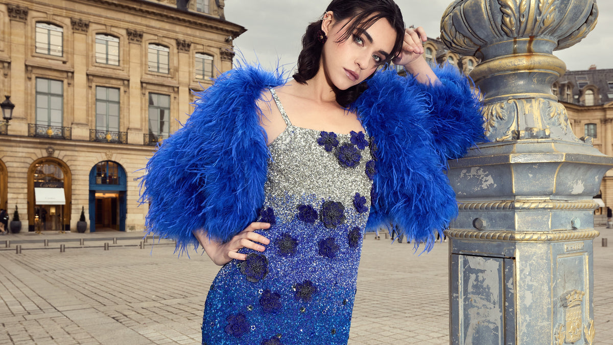 Model wearing a blue embellished gown with feathered sleeves beside a fountain in a city square.