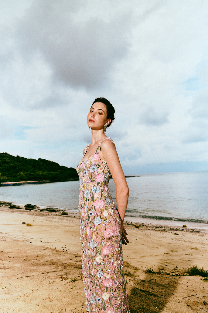 A woman stands on a sandy beach wearing a long, sleeveless floral dress with pink, peach, and cream tones. The background shows water, distant green hills, and a cloudy sky.