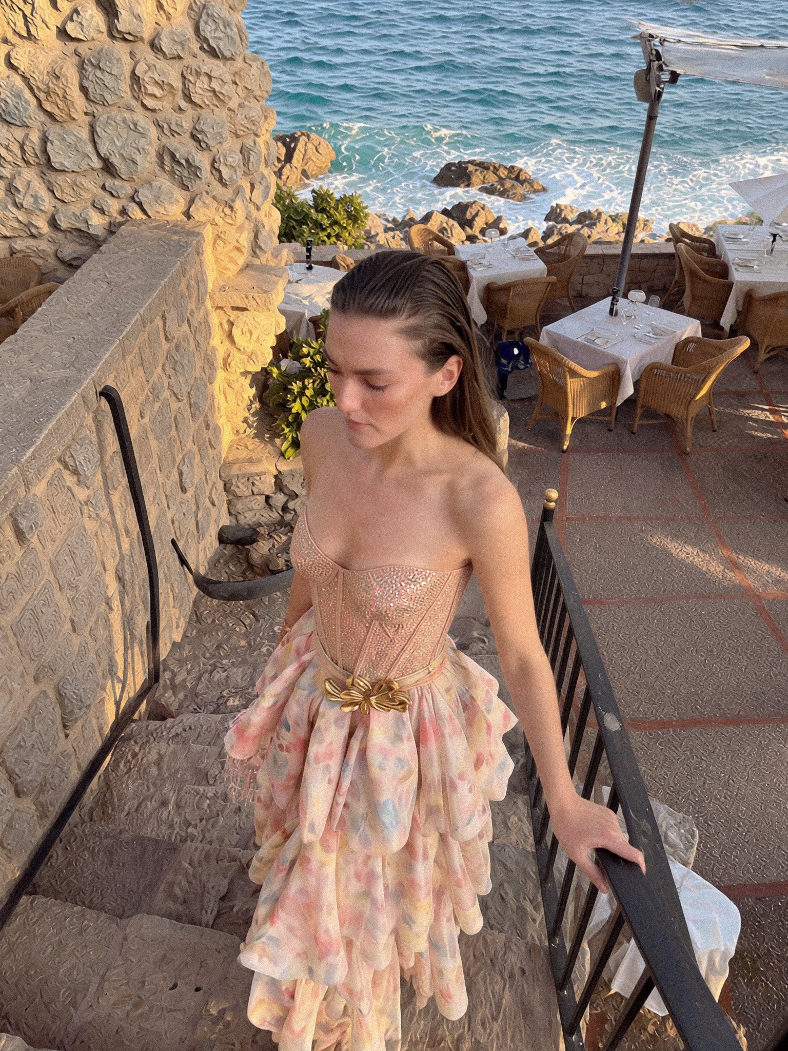 Woman in a floral dress standing on a stone pathway near a coastal restaurant.