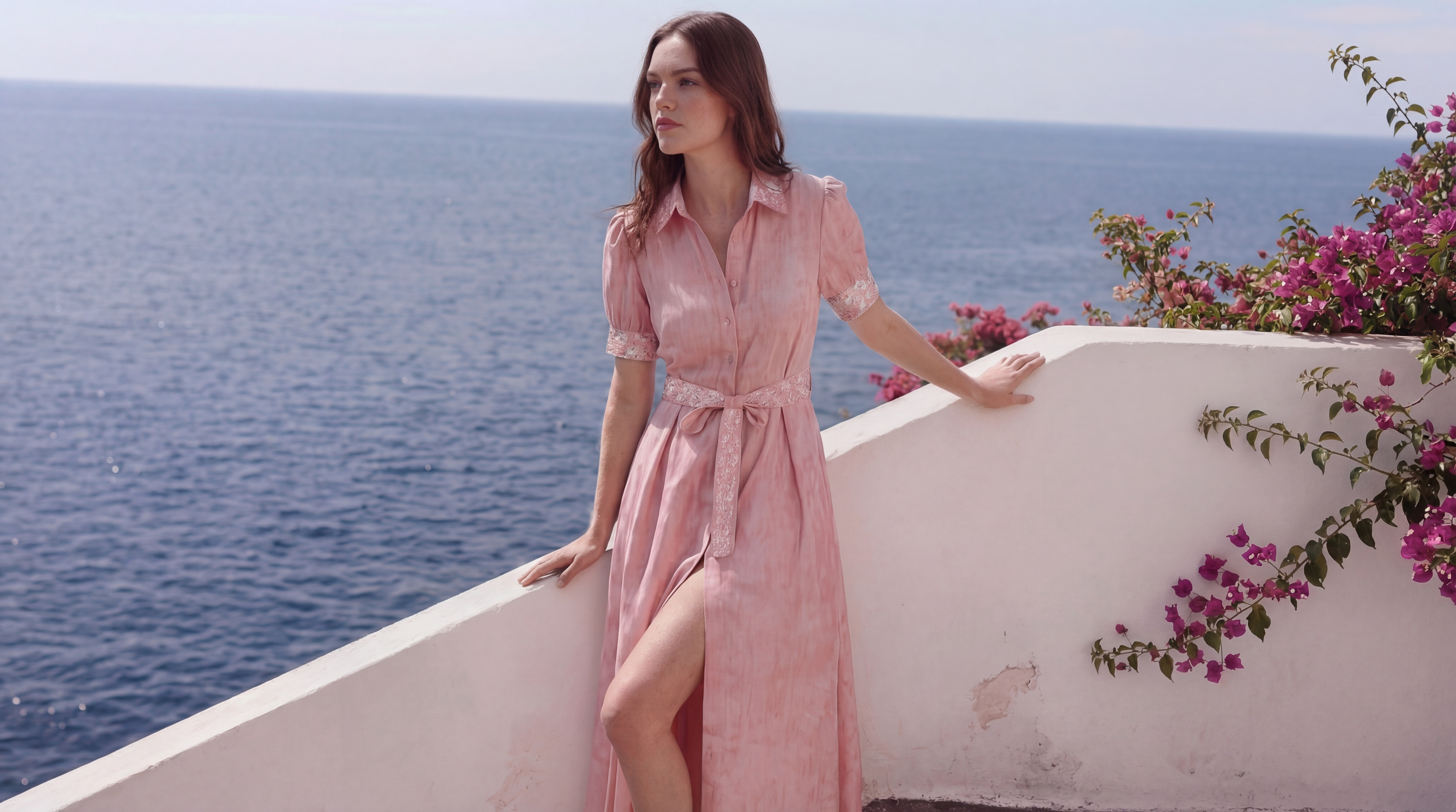 Woman in a pink dress standing on a balcony overlooking the ocean with flowers.