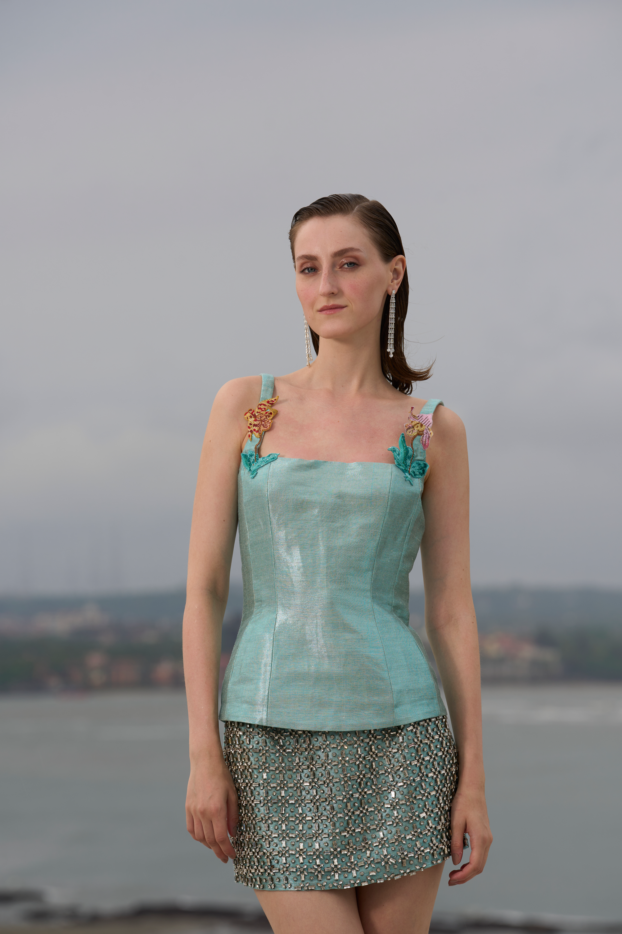 Woman wearing a light blue top and patterned skirt standing by a body of water.