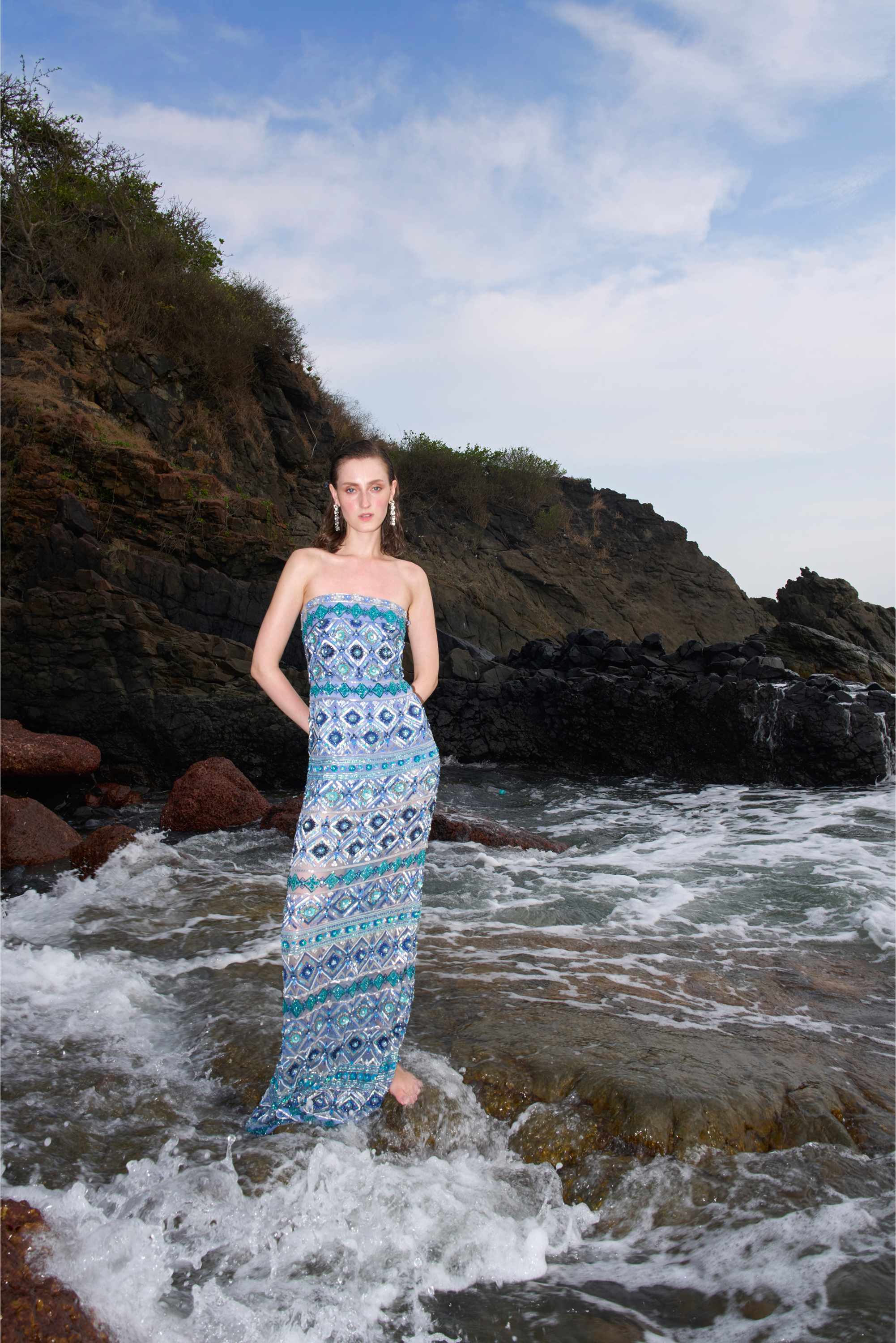 Woman in a blue strapless dress standing in the ocean with rocky cliffs and sky in the background