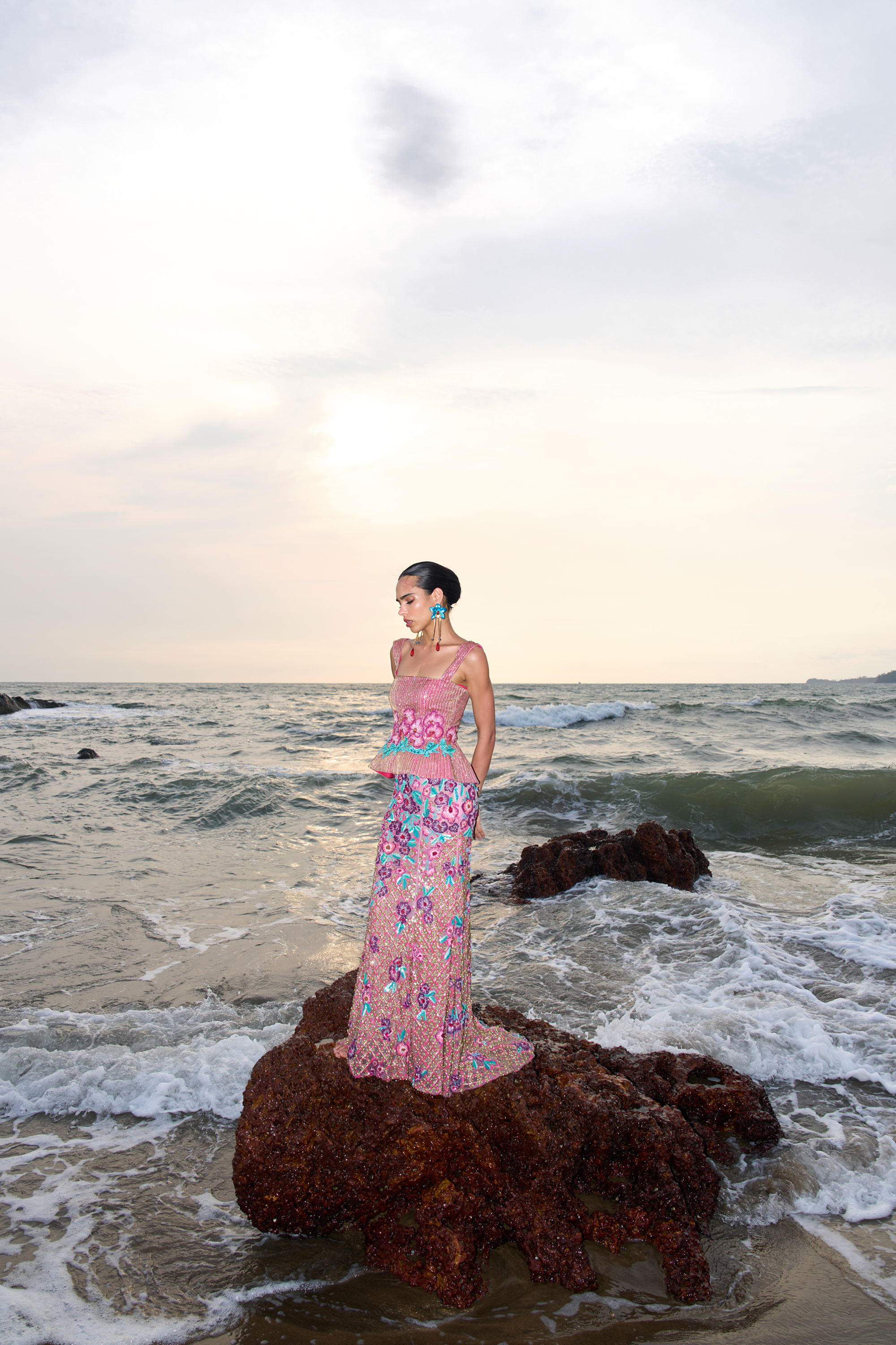 Woman in a pink floral dress standing on a rock in the ocean with a sunset sky.