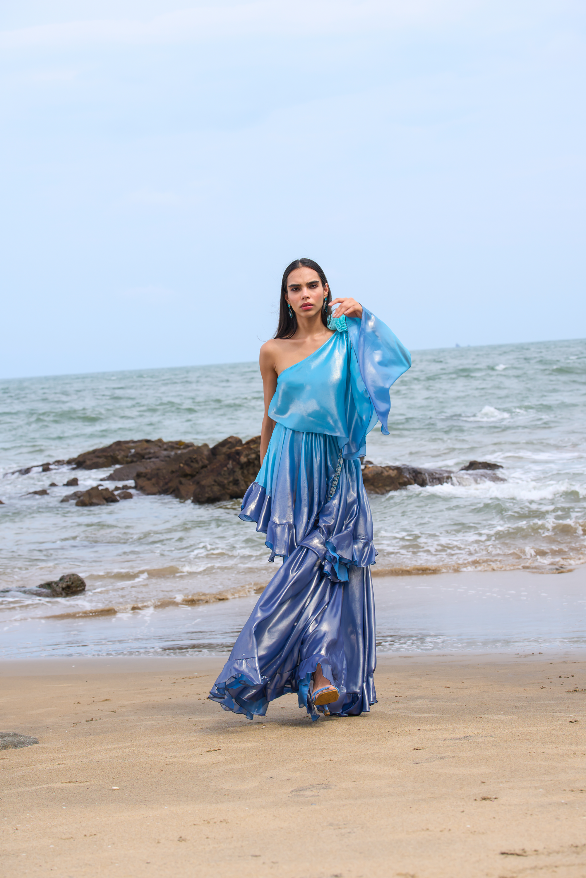 Woman in a blue dress standing on a beach with ocean and rocks in the background