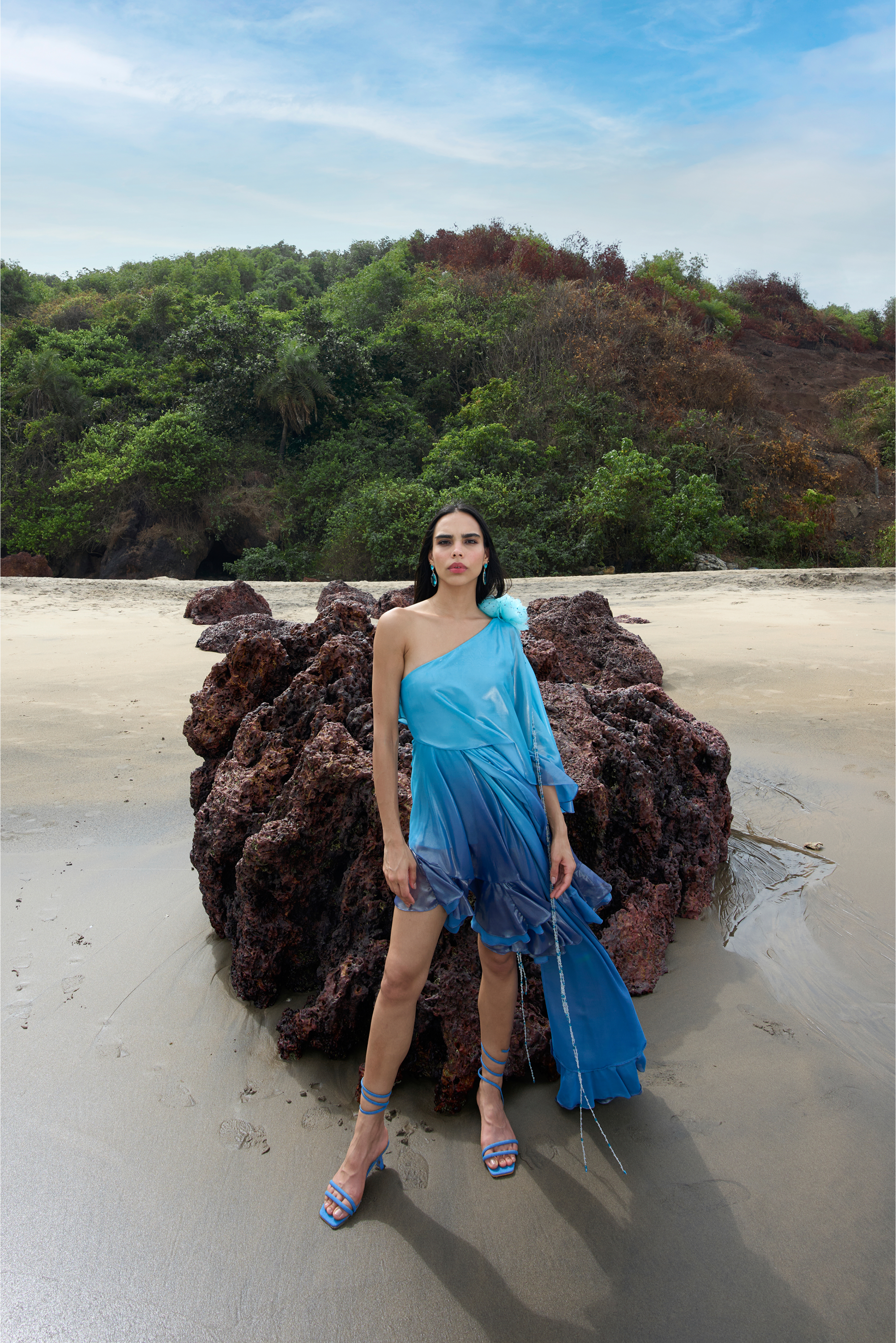 Woman in a blue dress standing on a beach with a rocky outcrop and greenery in the background
