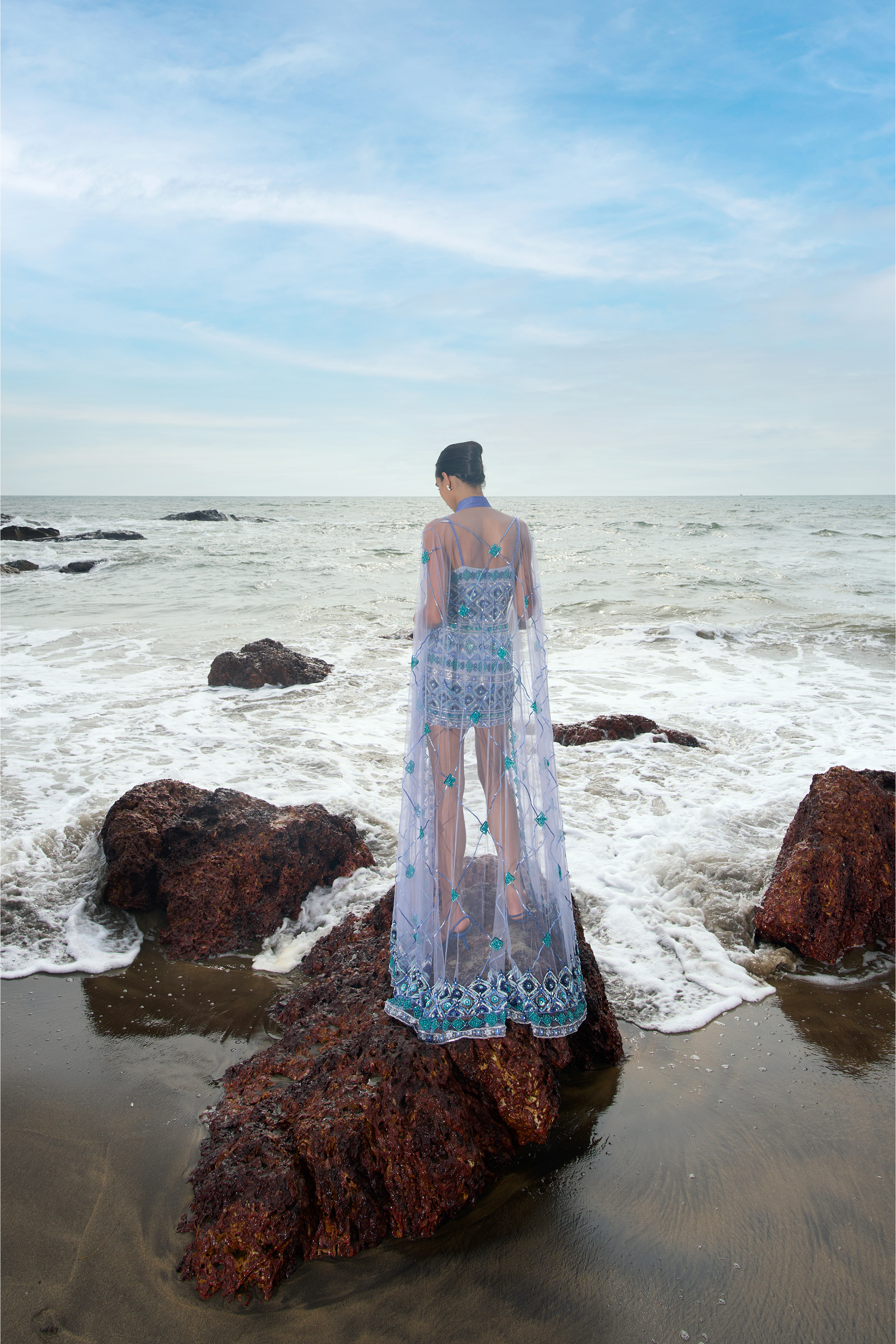 Person in a blue dress standing on a rocky beach with ocean waves.