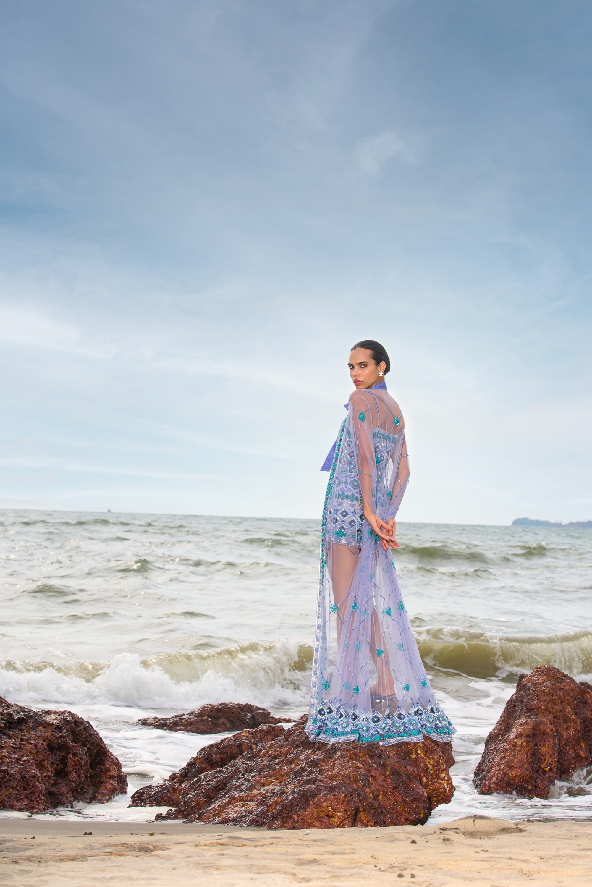 Person standing on a rocky beach with ocean waves and clear sky.