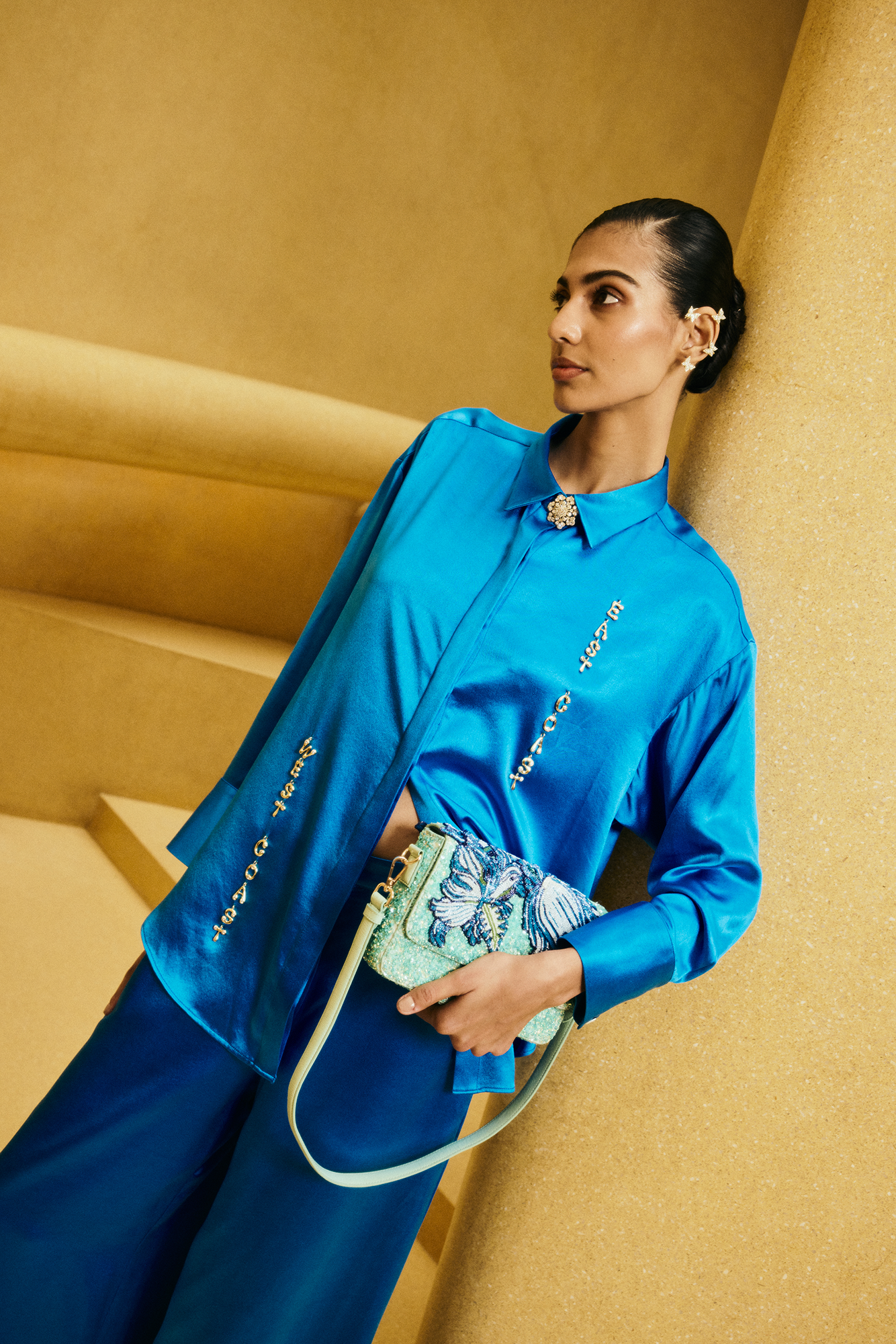 Woman in a blue outfit holding a patterned handbag against a beige wall.