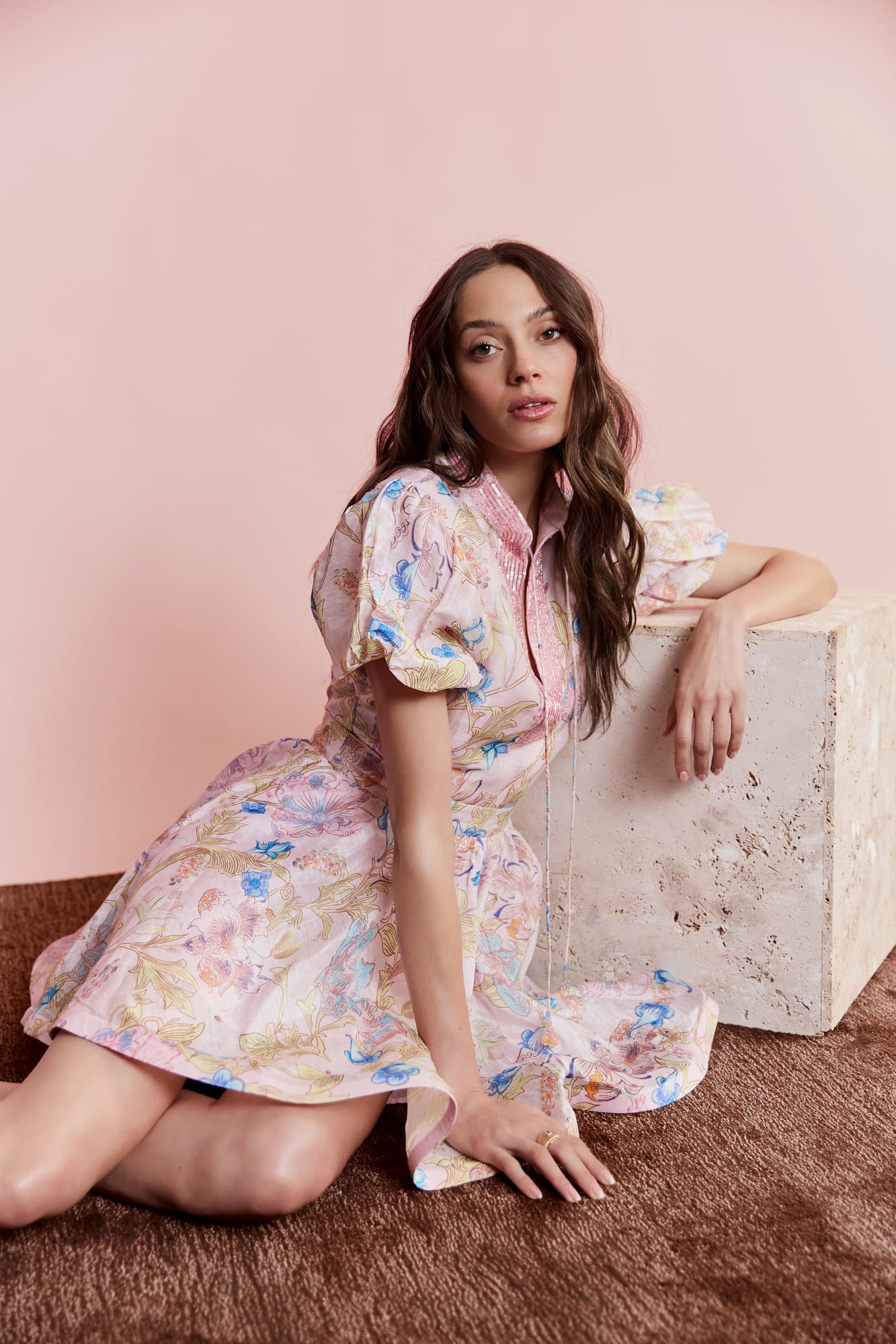 Woman in a floral dress sitting on a textured surface with a pink background