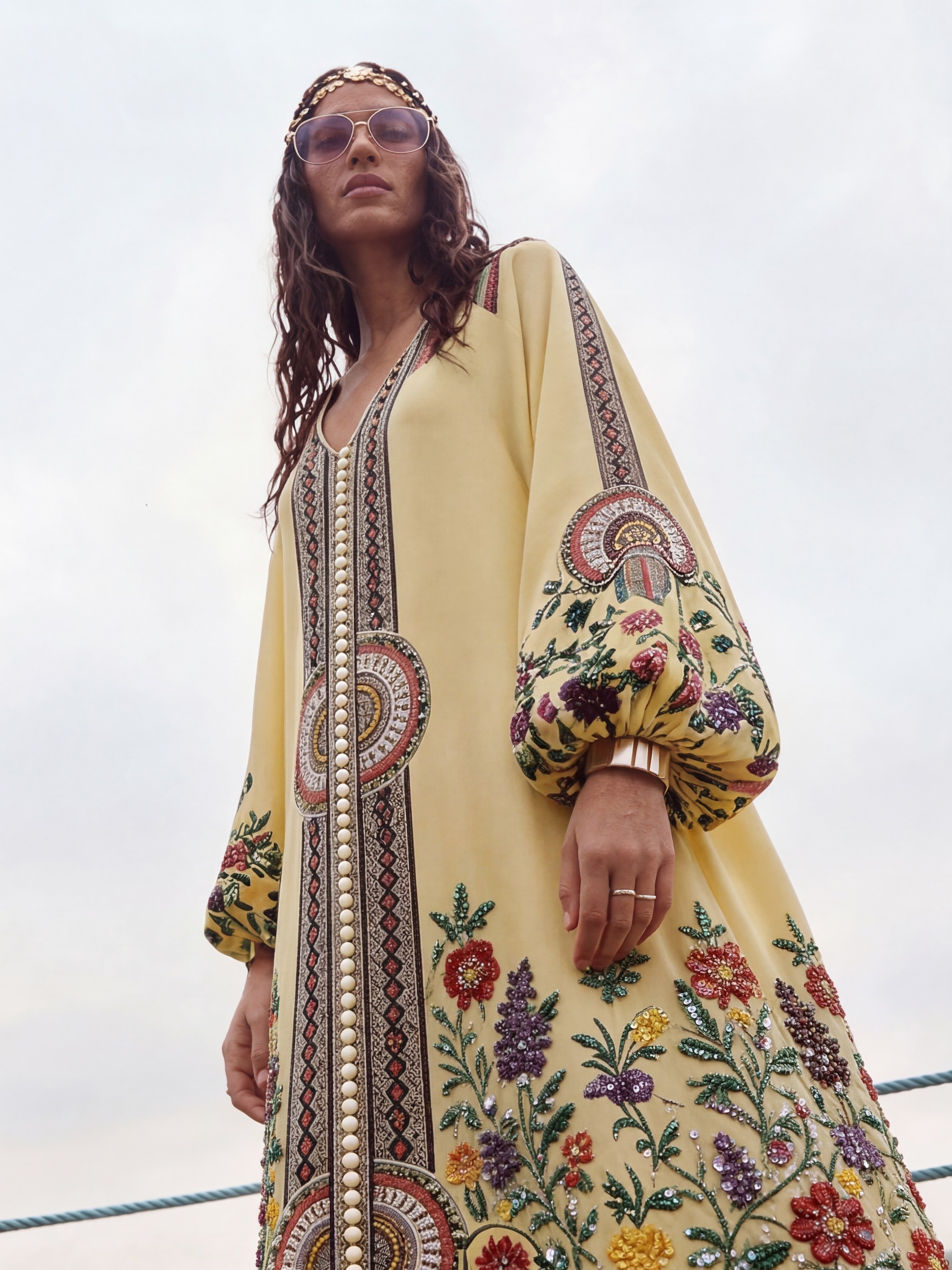 Woman wearing a yellow embroidered dress with floral patterns against a cloudy sky.