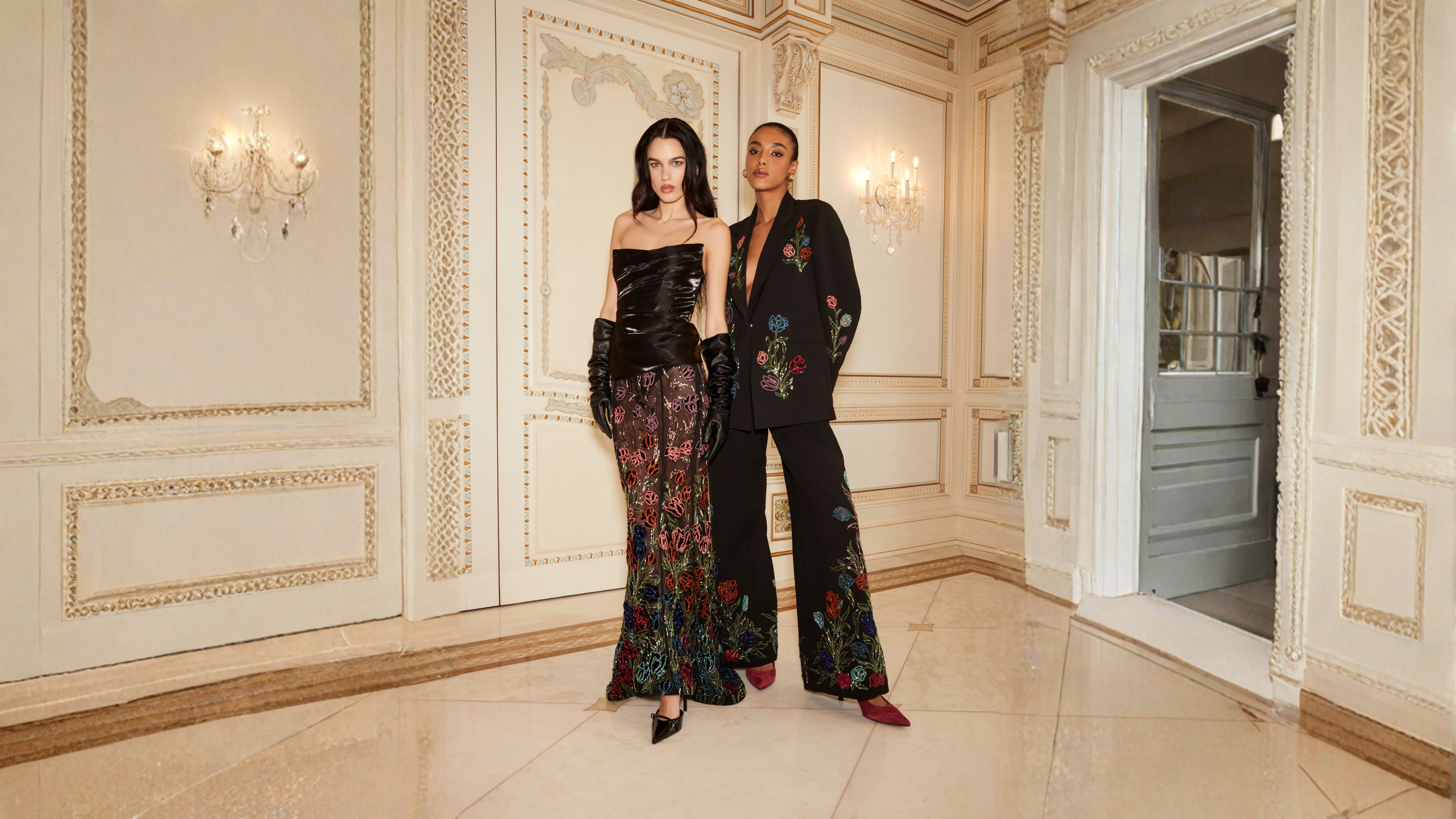 Two women in elegant outfits standing in a luxurious room with ornate walls.