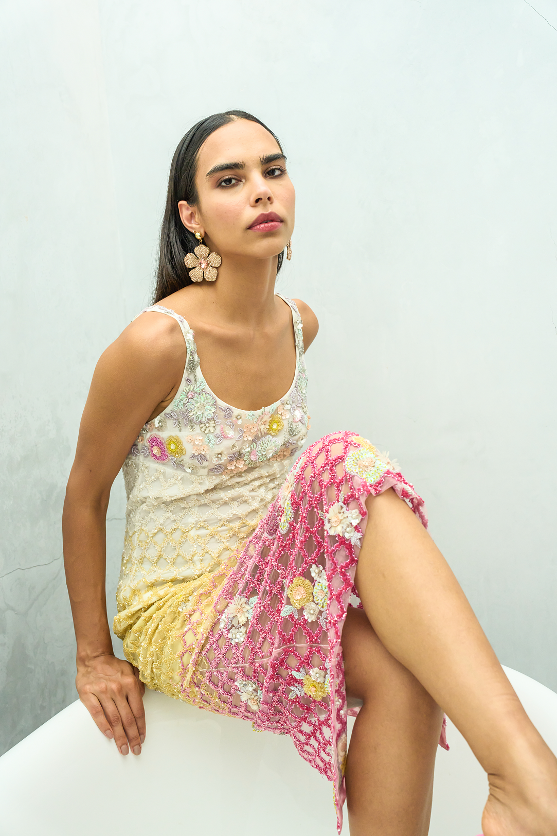 A woman models a sleeveless crochet lace dress with a gradient of cream, yellow, and pink, decorated with colorful floral embroidery. She is seated on a white curved surface against a light gray textured wall.