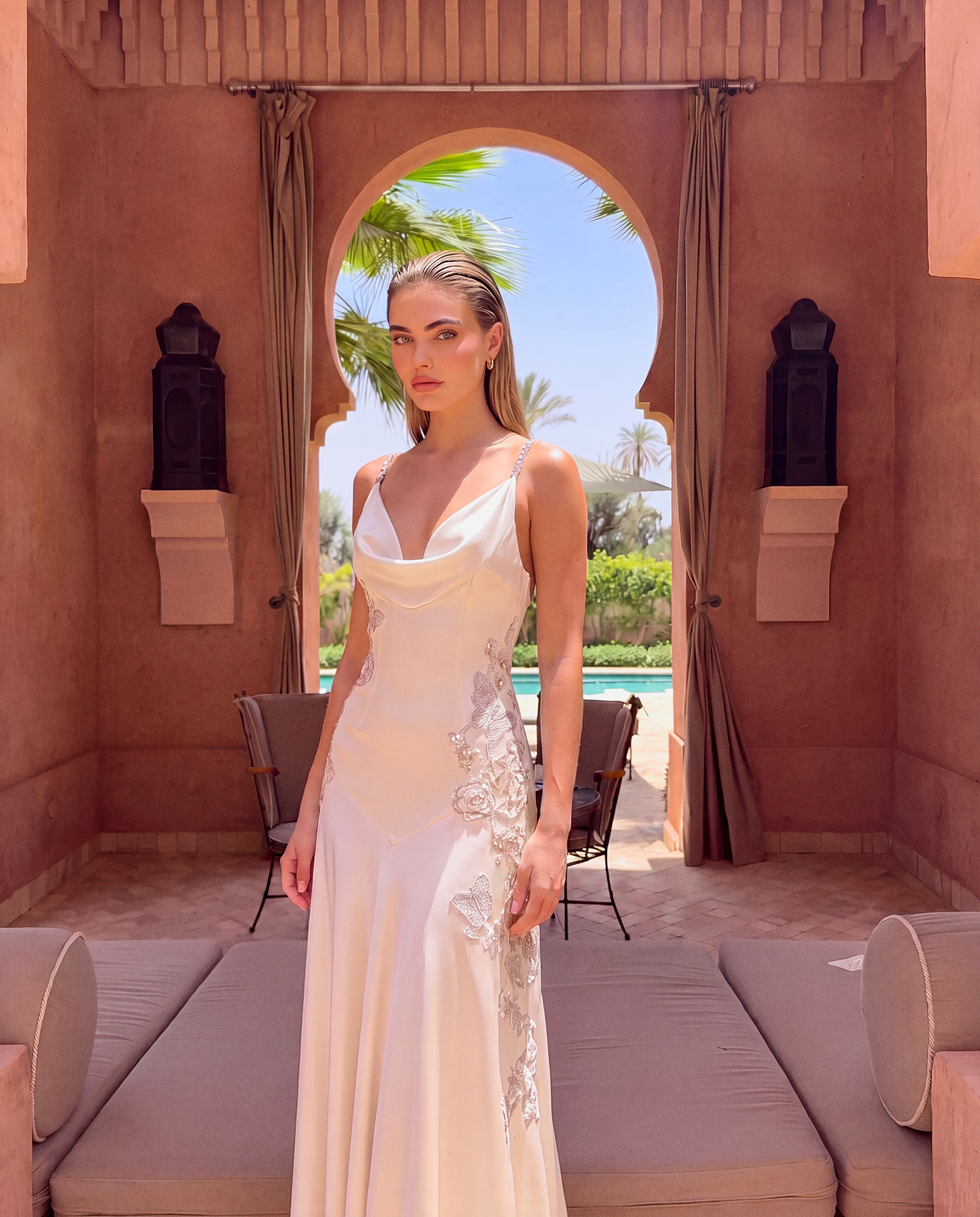A woman models a white sleeveless cowl-neck evening gown with silver lace applique on the side, standing in a luxurious terracotta-colored pavilion with an arched doorway leading to a poolside background.