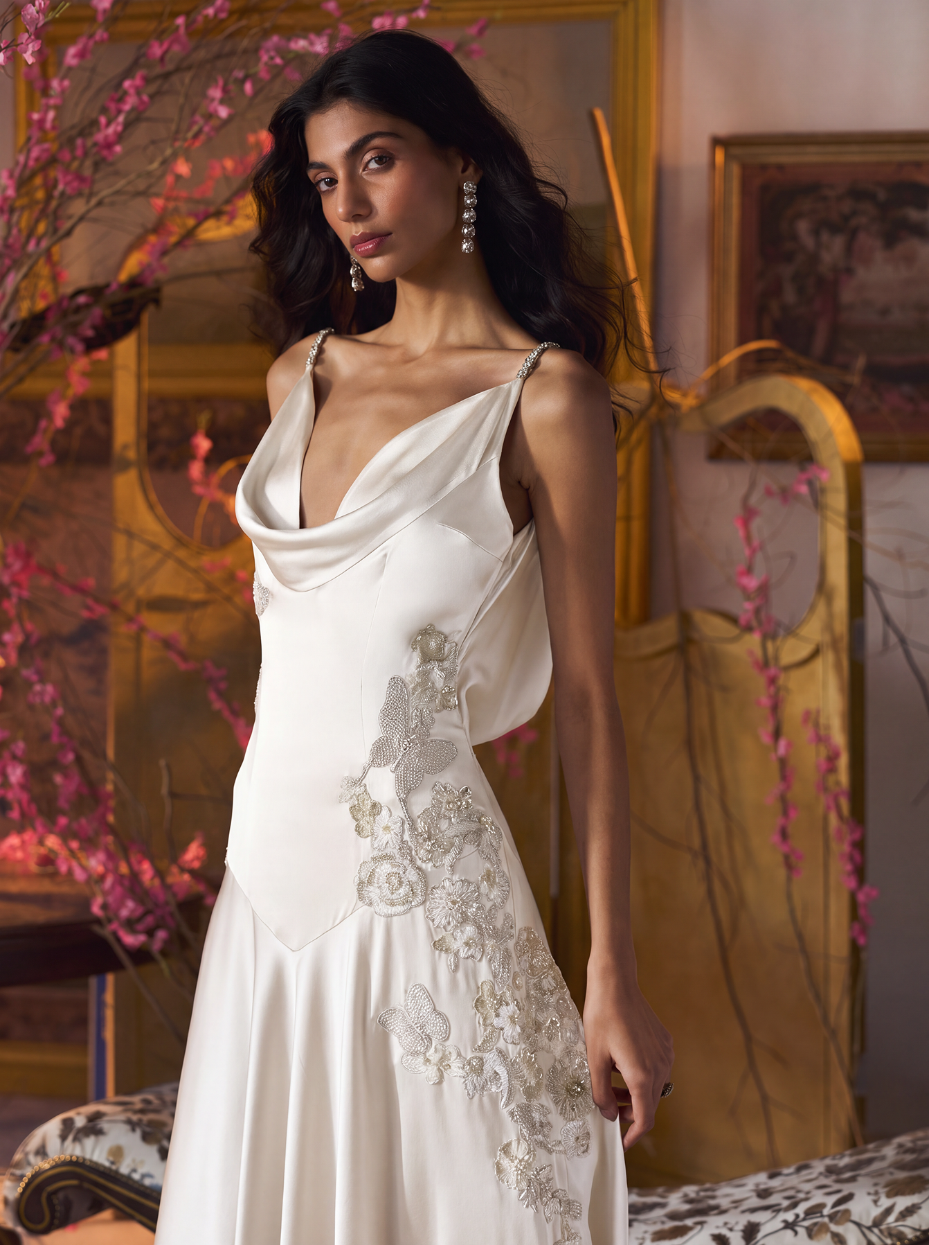 A woman models a white satin cowl-neck dress with spaghetti straps and floral embroidery on the bodice and skirt, posed in an elegant room with a gold headboard, pink blossoms, and vintage decor.