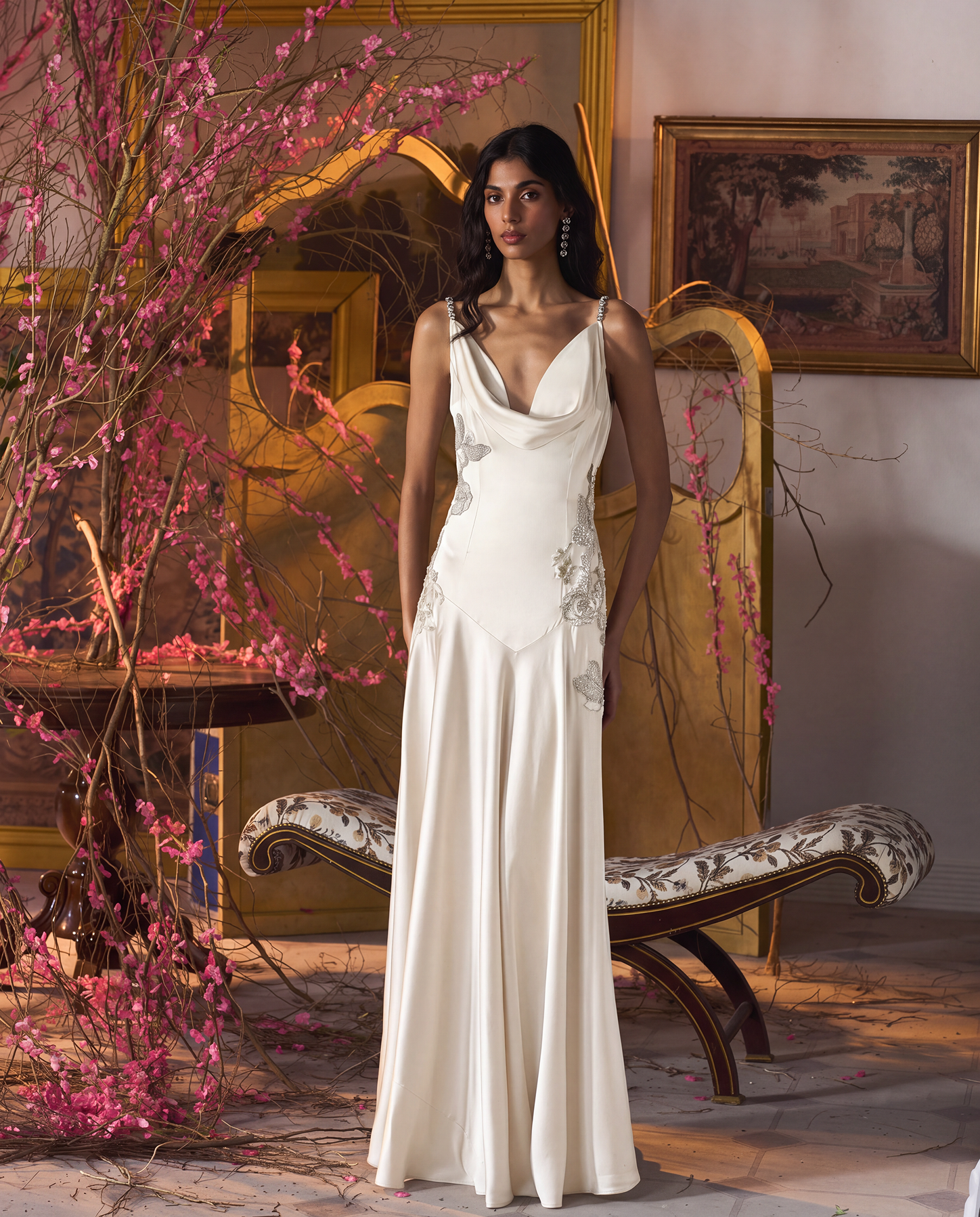 A woman stands indoors wearing a floor-length ivory gown with a draped neckline, fitted bodice, and flowing skirt. The room has warm neutral walls, ornate gold furniture, framed artwork, and pink flowering branches in the foreground.
