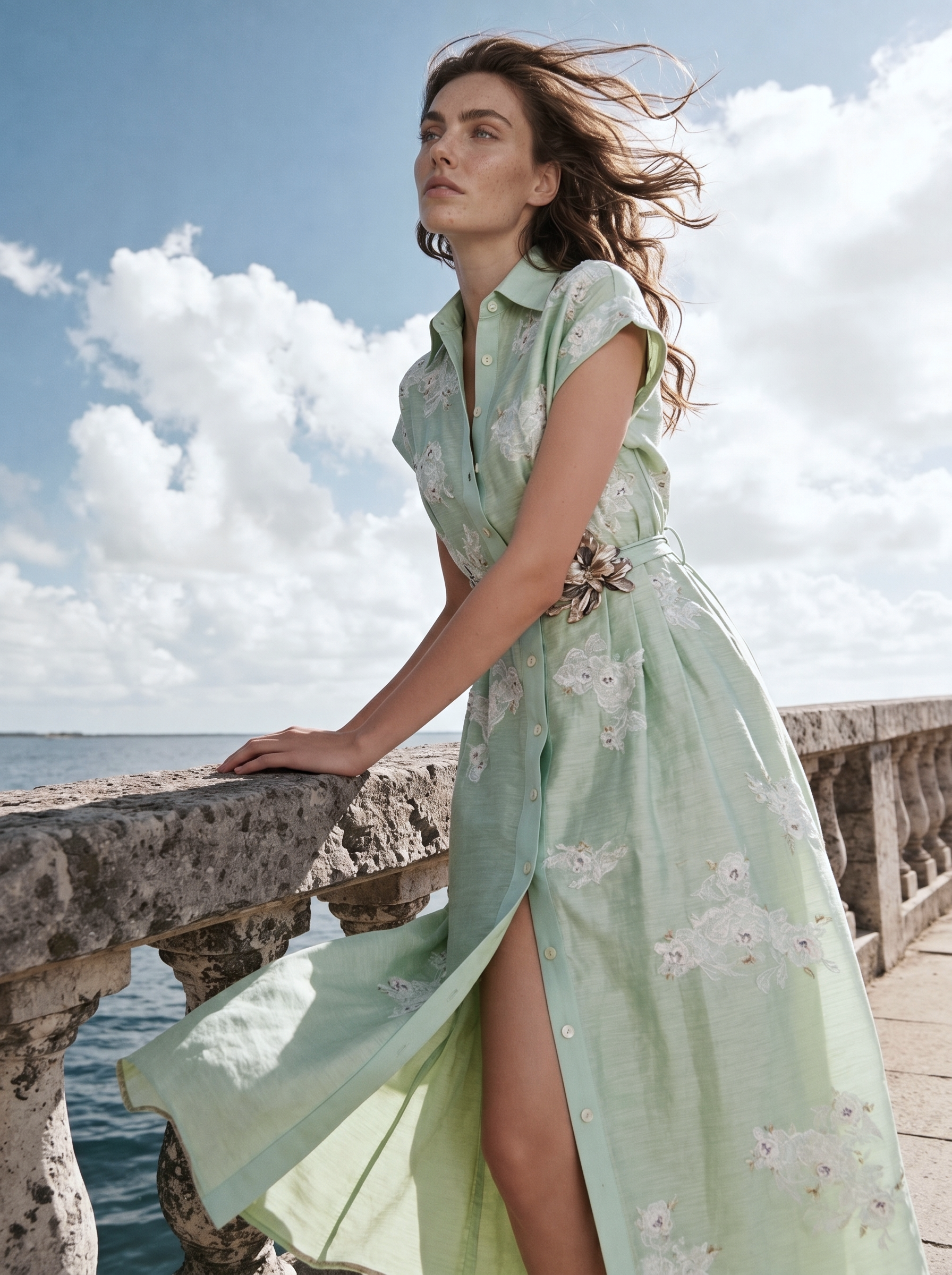 Woman in a light green floral dress standing by a waterfront with a cloudy sky.