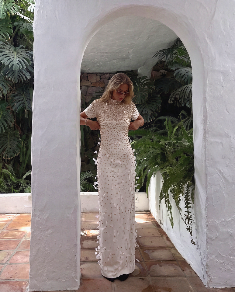 A woman wearing a long, fitted cream dress with textured floral appliqués stands inside a white stucco archway surrounded by lush green tropical plants.
