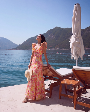 A woman wearing a pink and yellow floral maxi dress stands on a sunlit patio by the sea, holding a white hat, with wooden lounge chairs, a closed umbrella, and mountains in the background.