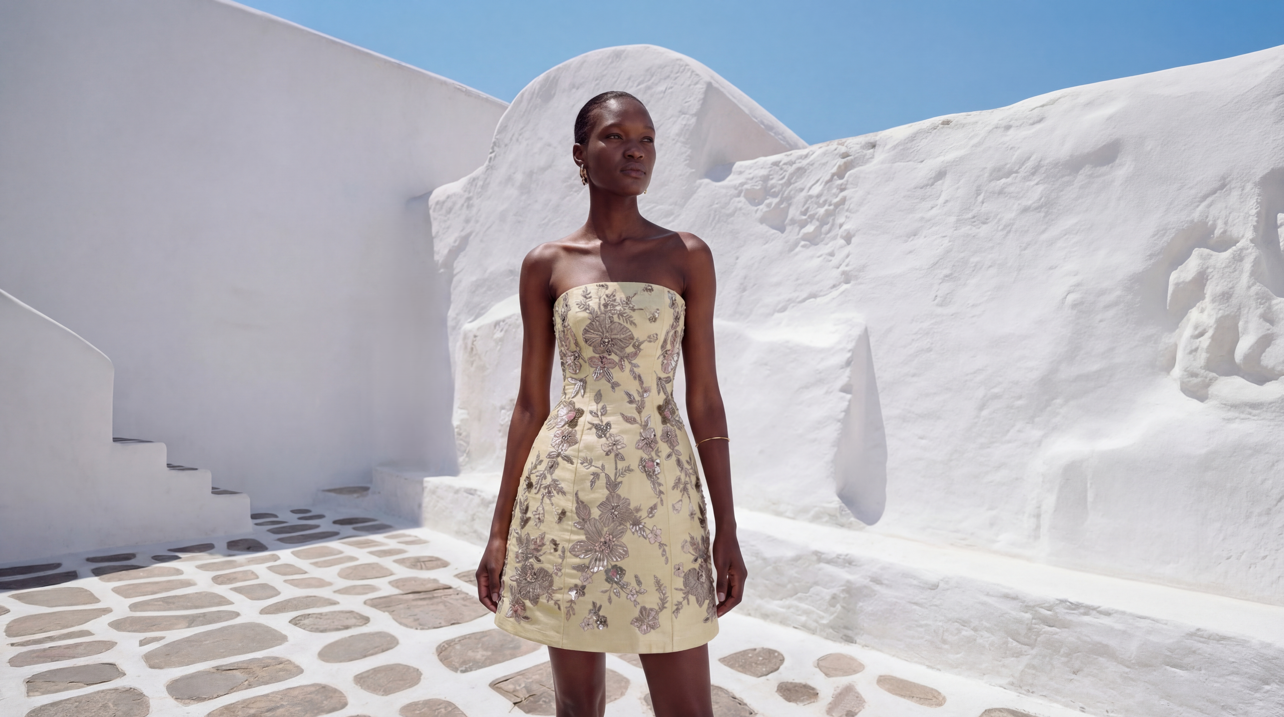 Woman in a floral dress standing in a sunlit, white-walled outdoor setting.